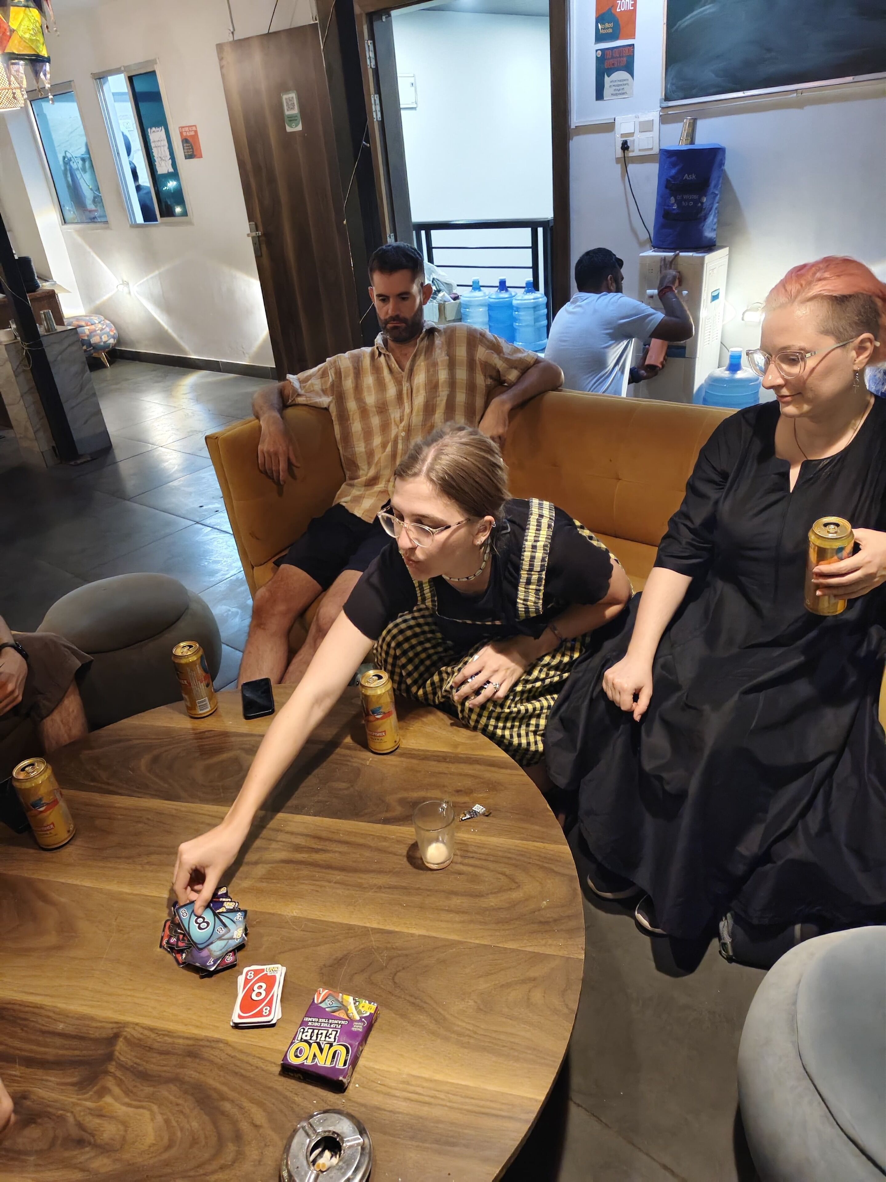 A group of people seated around a wooden table with snacks and drinks, in a room with a large window and a door.