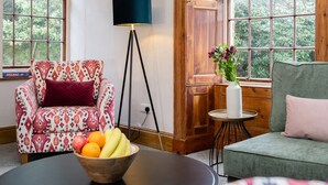 A room with a patterned armchair, a green sofa, a wooden door, and a window with a view of greenery. - Watermouth Castle, Rhododendron Apartment (Ilfracombe)