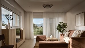Ein modernes Wohnzimmer mit Sofa, Hocker und Couchtisch. Ein Fernseher steht auf einem Holzgestell, eine Zimmerpflanze sorgt für Grün und durch große Fenster eröffnet sich der Blick ins Freie.
