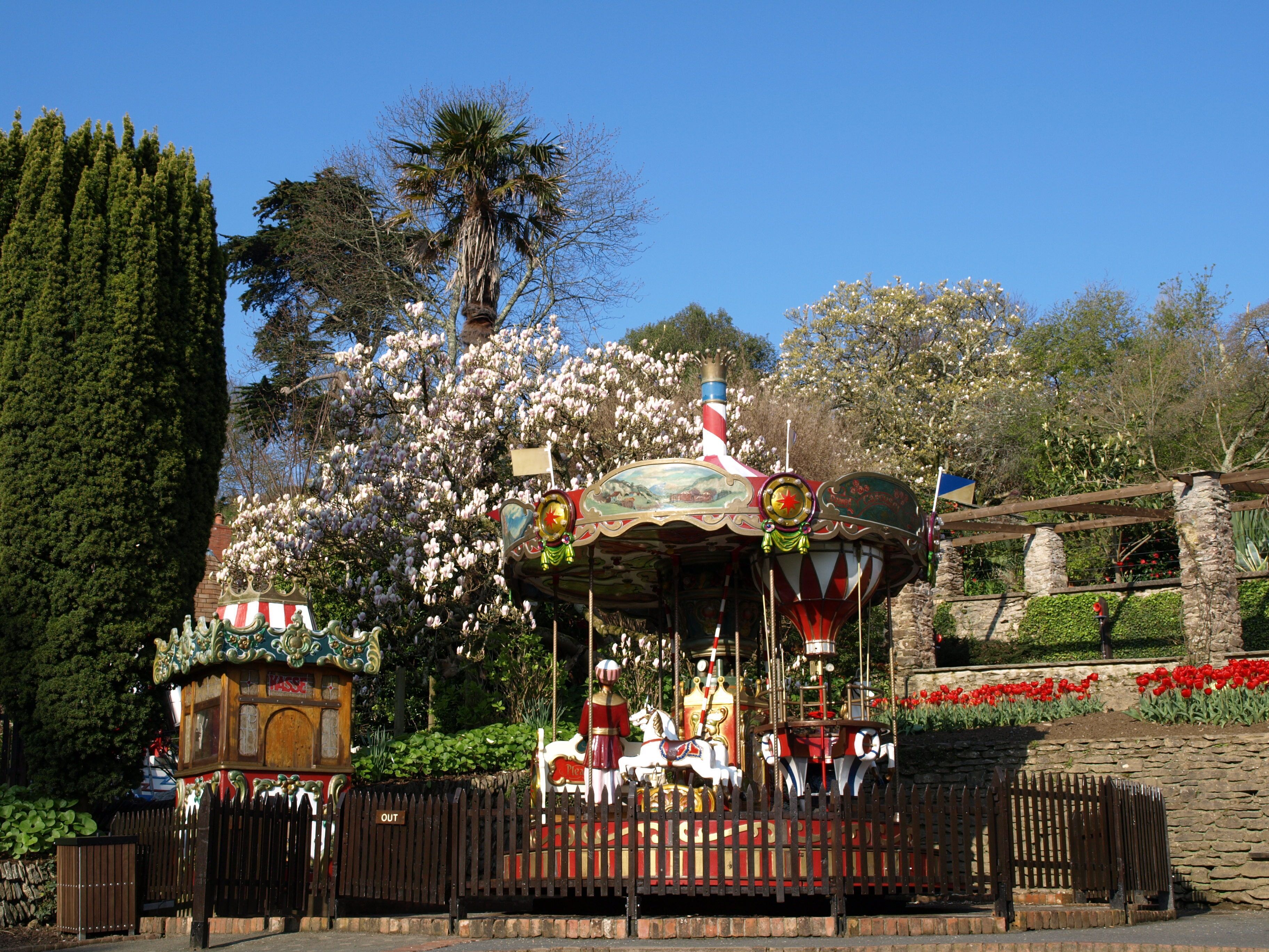 A carousel with ornate decorations and a figure on top, surrounded by blooming trees and a stone wall with red flowers.