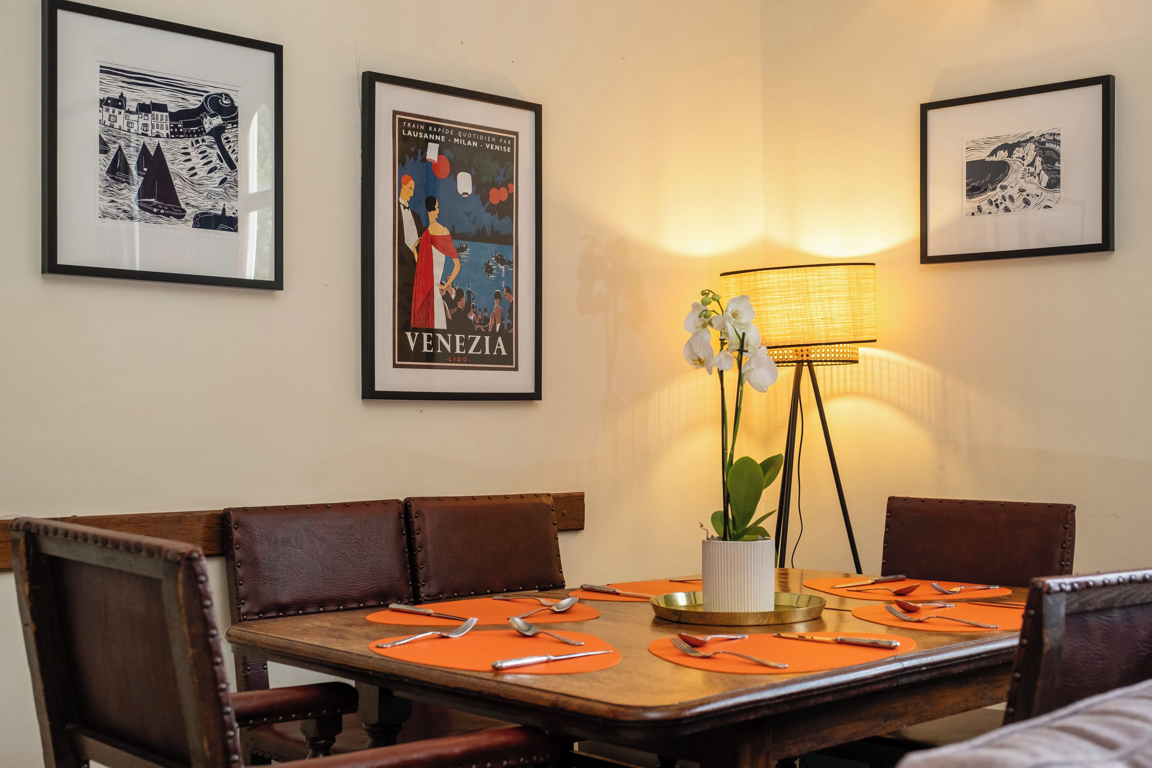 Een eetruimte met een houten tafel gedekt voor vier personen, afbeeldingen aan de muur en een vloerlamp.