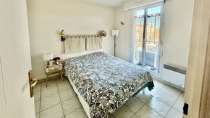 A bedroom with a bed, a nightstand, a lamp, a window with a view, and a radiator. - Beautiful 65 m² flat (Saint-Raphaël)
