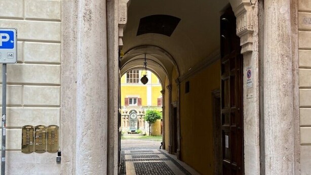 Um corredor estreito com arquitetura clássica, incluindo colunas e teto em arco, que leva à entrada de um edifício.