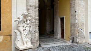 Corredor de arquitetura clássica com uma estátua de mármore de uma mulher e criança, portas em arco e piso de pedra.