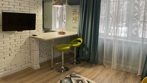 A modern kitchen with a brick wall, a flat-screen TV, a wooden countertop, two green bar stools, and a large window with sheer curtains. - NicePlace in the Old Center Almaty (Almaty)
