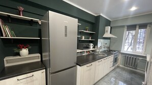 A modern kitchen with a stainless steel refrigerator, a microwave, a sink, and a stove. There are wooden cabinets and a window with a view of the outside. - NicePlace in the Old Center Almaty (Almaty)