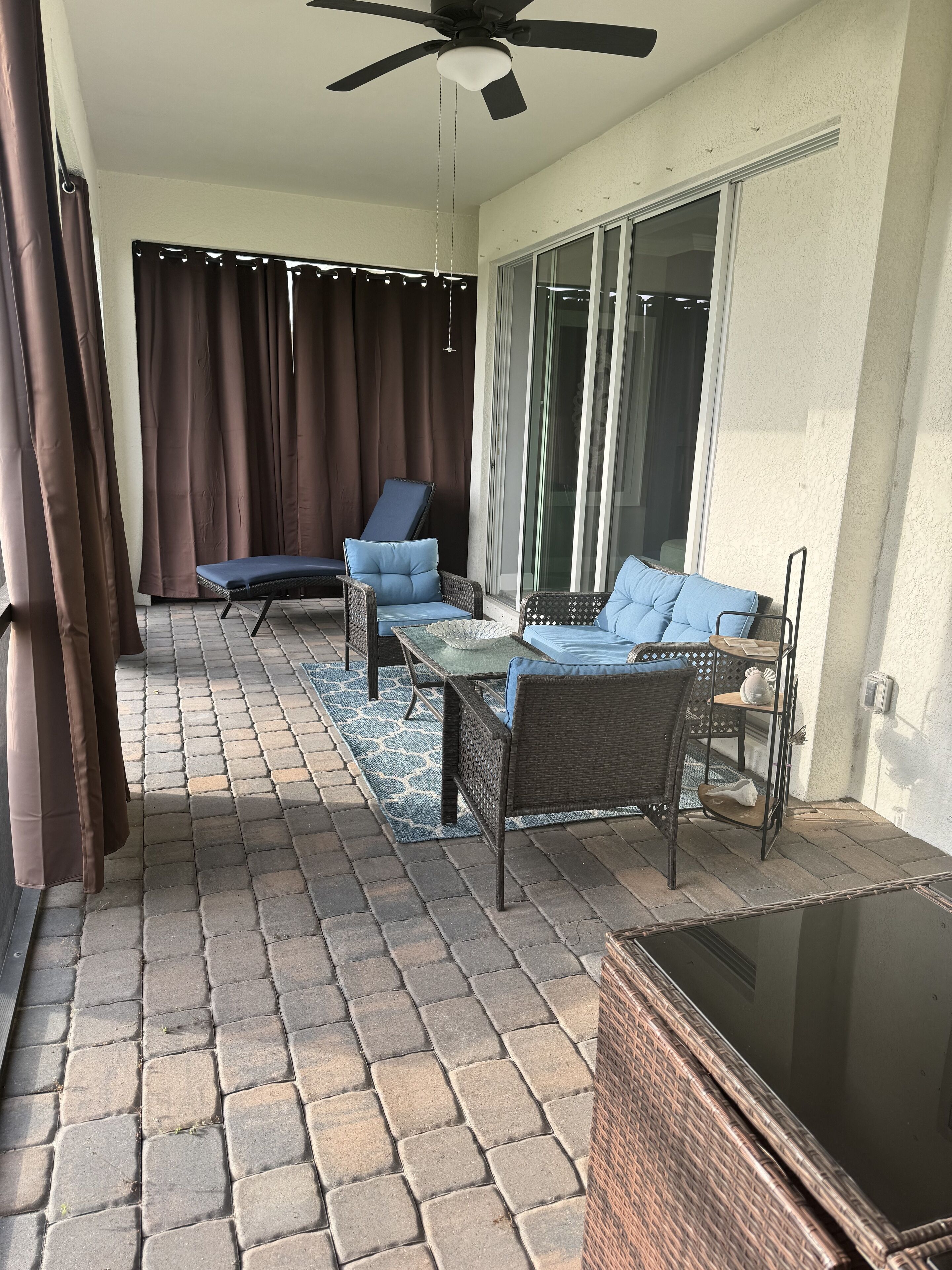 A covered outdoor area with a ceiling fan, two blue cushioned chairs, a small table, and a wicker chair.