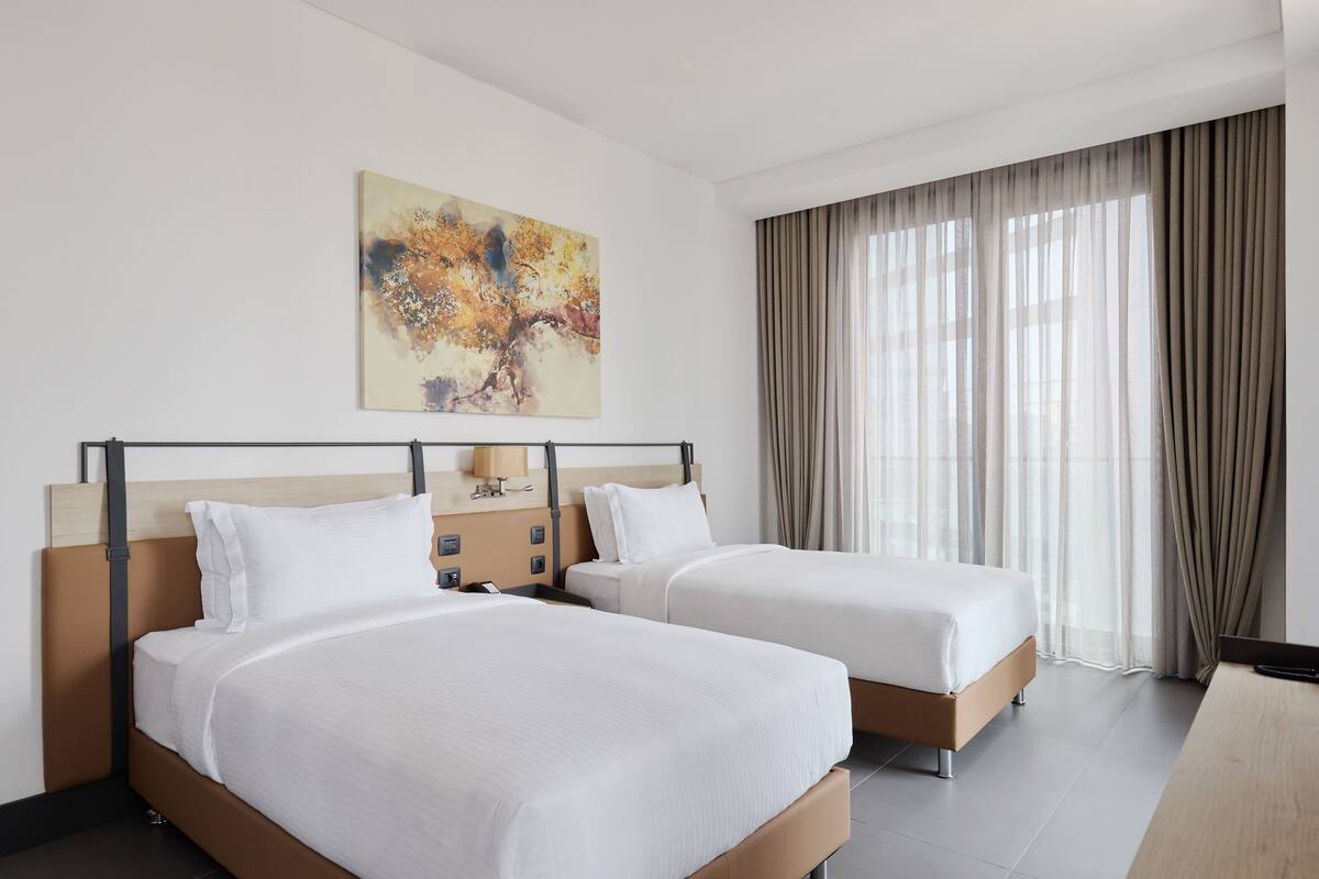 A hotel room with two beds, a painting on the wall, and a large window with curtains.