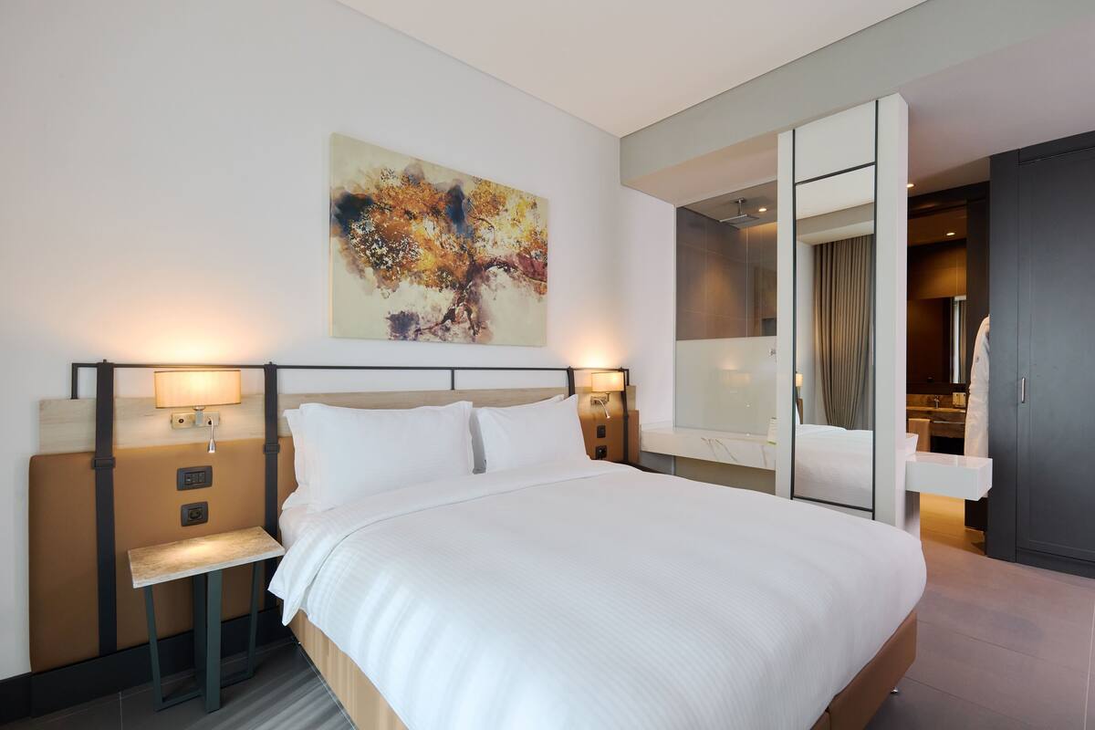 A modern hotel room with a large bed, bedside lamps, a nightstand, a painting on the wall, and a bathroom visible through an open door.