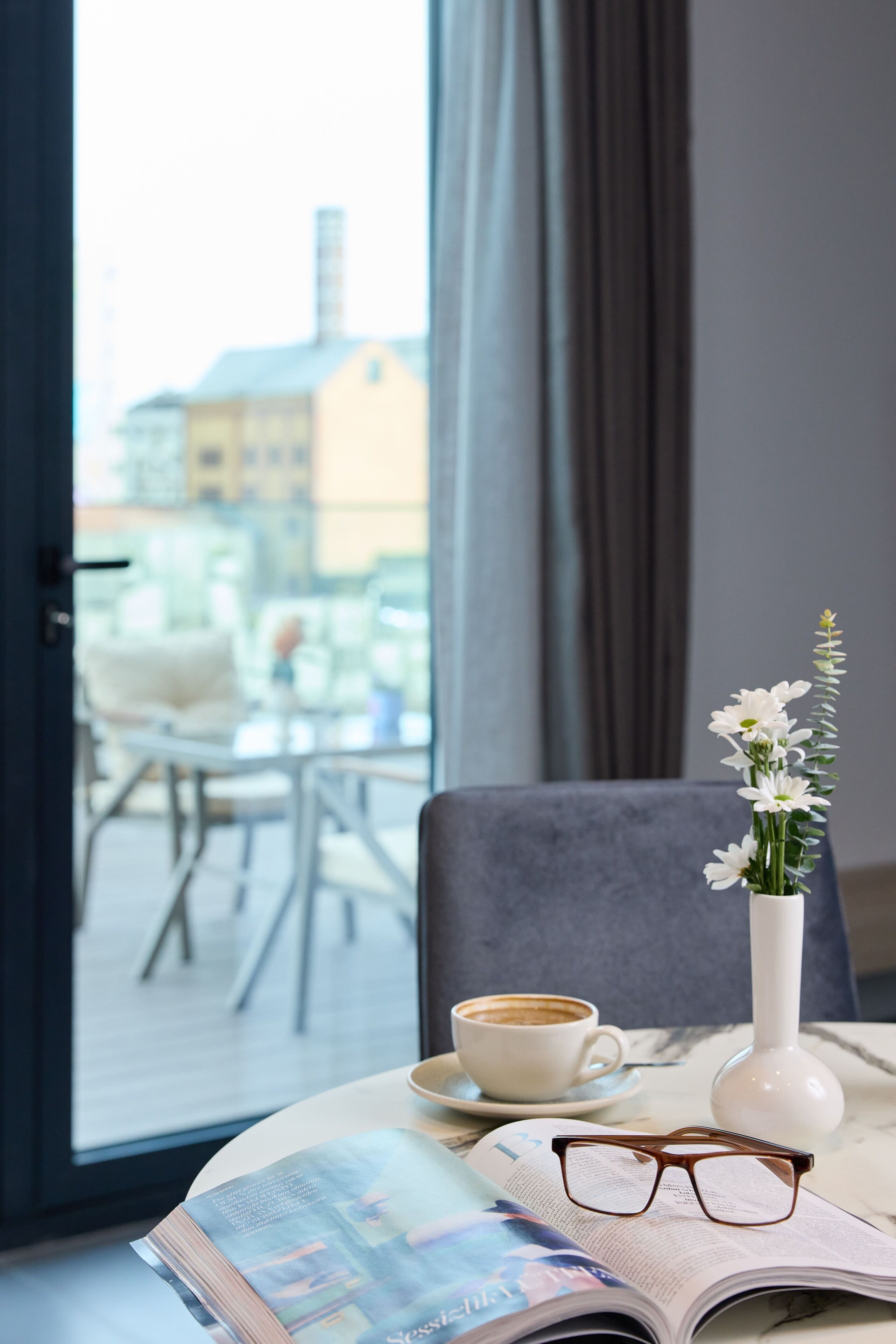 Een kamer met een tafel waarop een kop koffie, een vaas met bloemen, een paar brillen en een open tijdschrift staan.