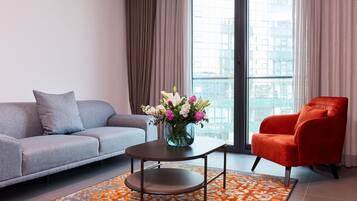 Un salon moderne avec un canapé gris, un fauteuil rouge, une table basse ornée d’un vase de fleurs et une grande fenêtre avec des rideaux.