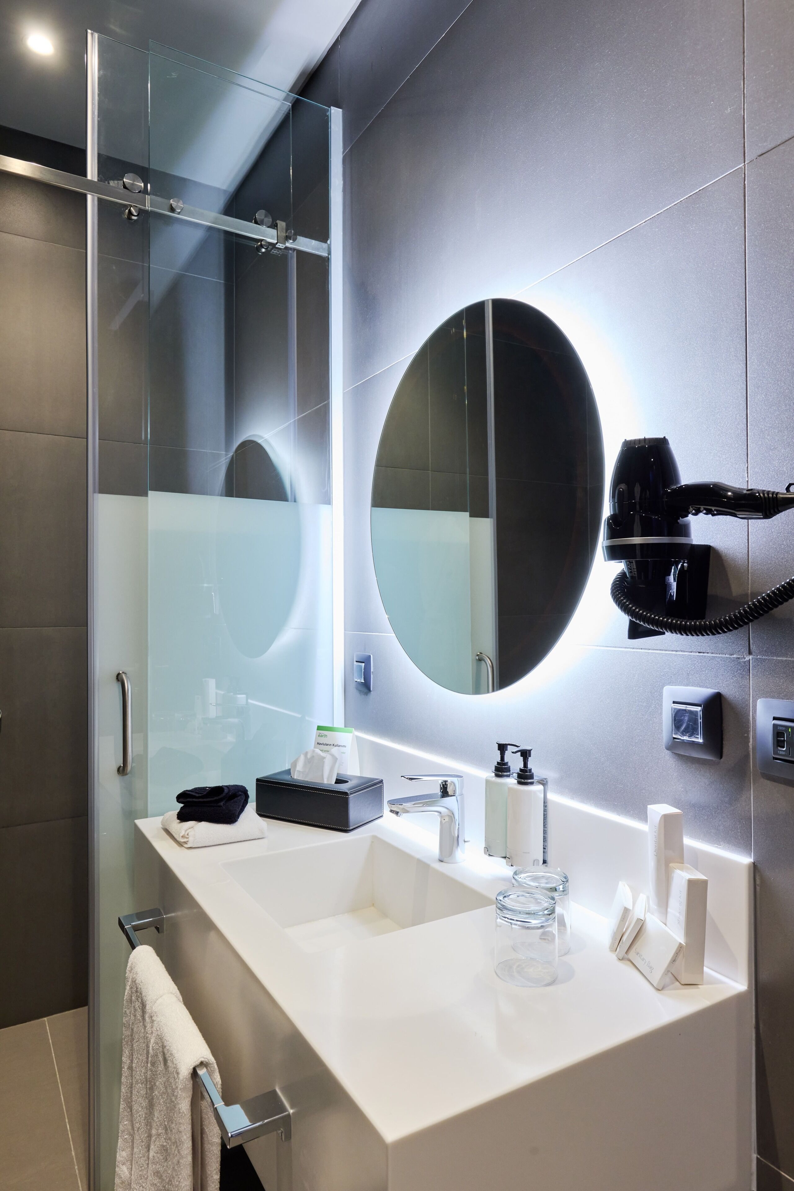 Ein modernes Badezimmer mit einer in Glas abgeschirmten Dusche, einem großen Spiegel und einem an der Wand befestigten Föhn.