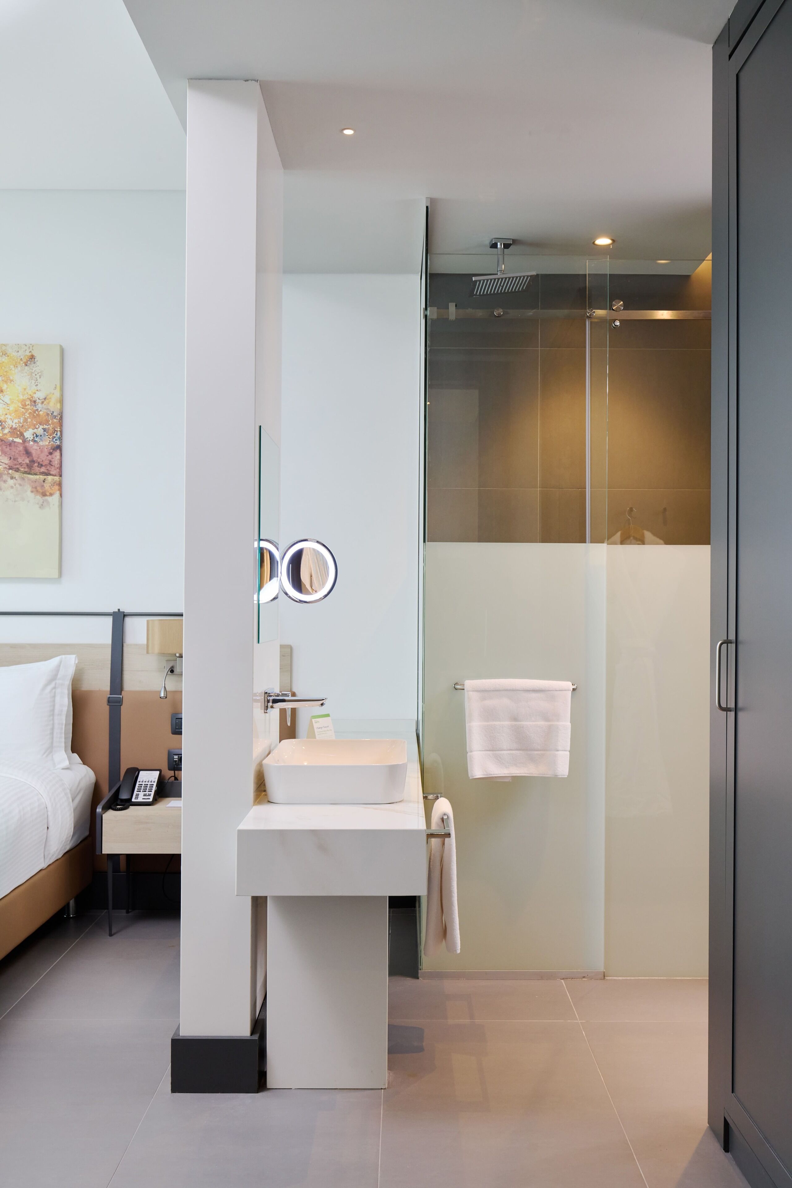 A modern bathroom with a glass shower enclosure, a white sink, and a bedside table with a phone.