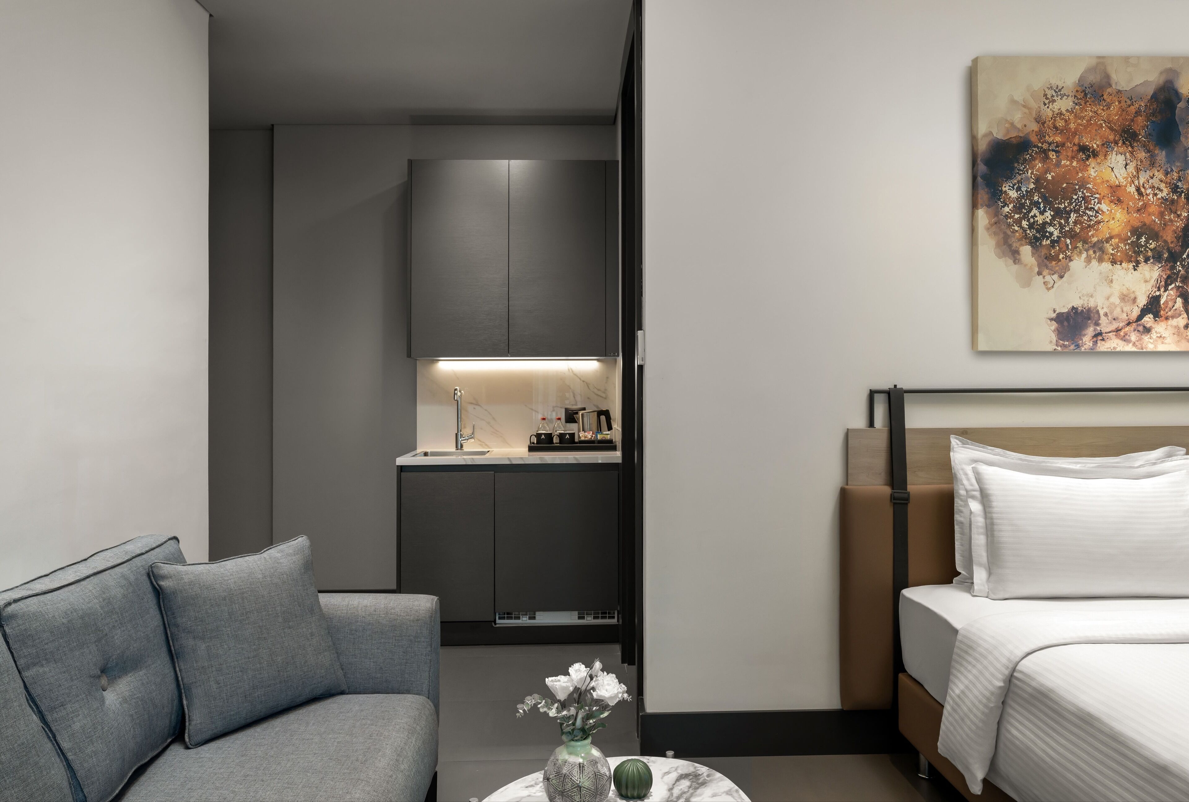 A modern hotel room with a grey sofa, a round coffee table with a vase, a bed with white linens, a small kitchen area, and a painting on the wall.