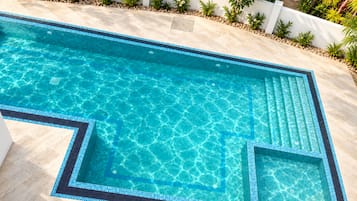 Una piscina rectangular con agua azul cristalina, rodeada por un muro blanco y una diversidad de plantas verdes.