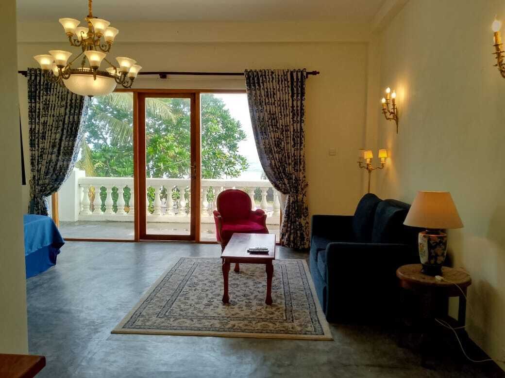 A living room with a red chair, a blue sofa, a coffee table, and a chandelier. There is a balcony with curtains and a view of trees.