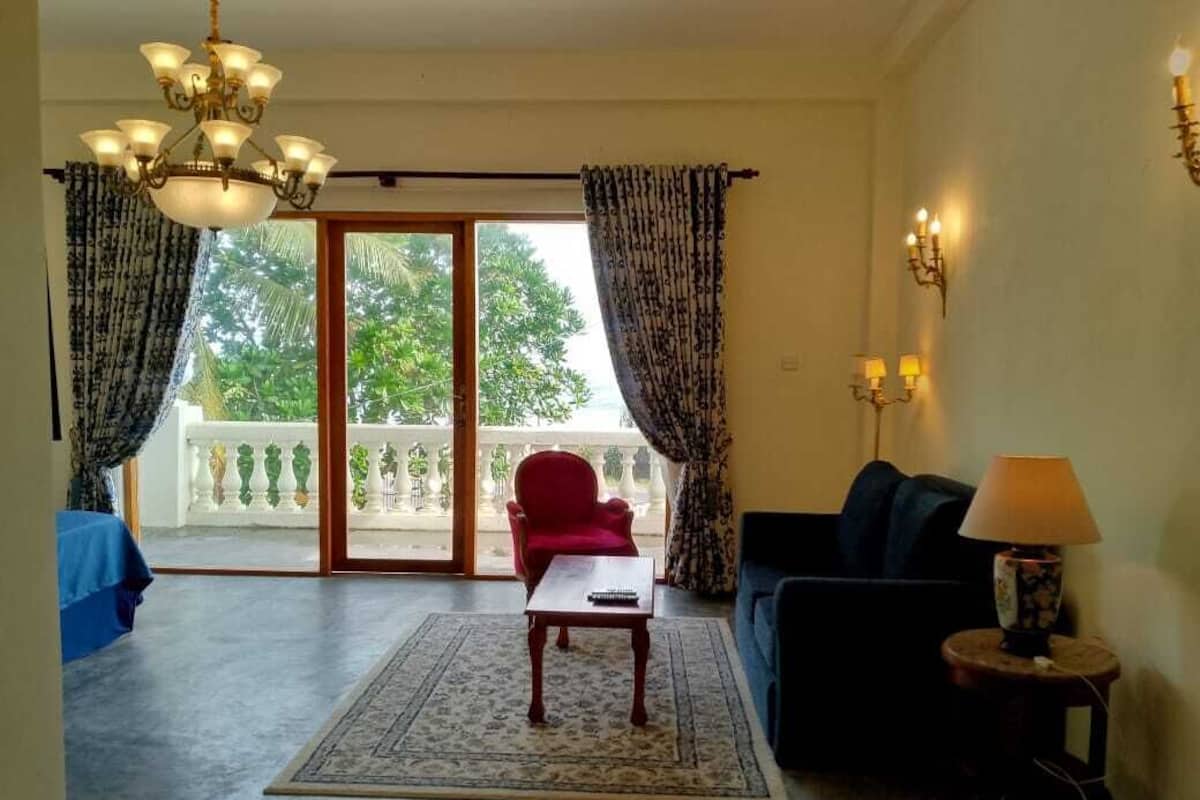 Sala de estar com poltrona vermelha, sofá azul, mesa de centro e lustre. Há uma varanda com cortinas e vista para árvores.