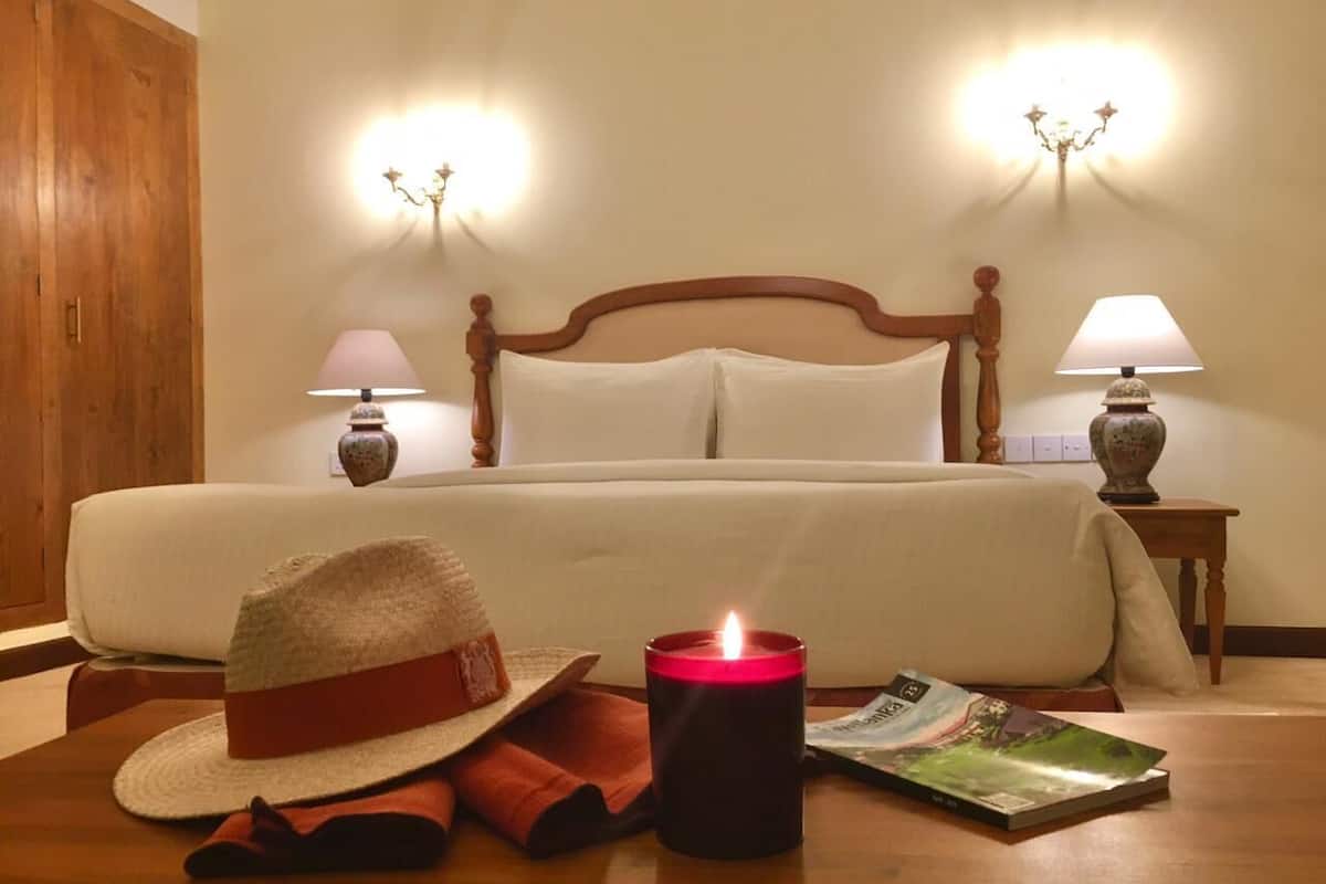 Quarto de hotel com cama, abajures, uma vela, um chapéu e uma revista no chão.