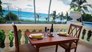 A wooden table set for two with plates, glasses, and a bottle of wine, overlooking a beach with palm trees and the ocean. - Samsara (Boossa)