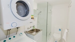 Une salle de bain de taille réduite, équipée d’un lave-linge à chargement frontal, d’une douche à parois de verre et de toilettes classiques.