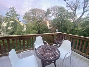 A wooden deck with a round table and four white chairs, surrounded by trees and a building in the background. - Oceanside, Kid-Friendly Emerald Isle Home w/ Hot Tub and 2 Kitchens (Emerald Isle)