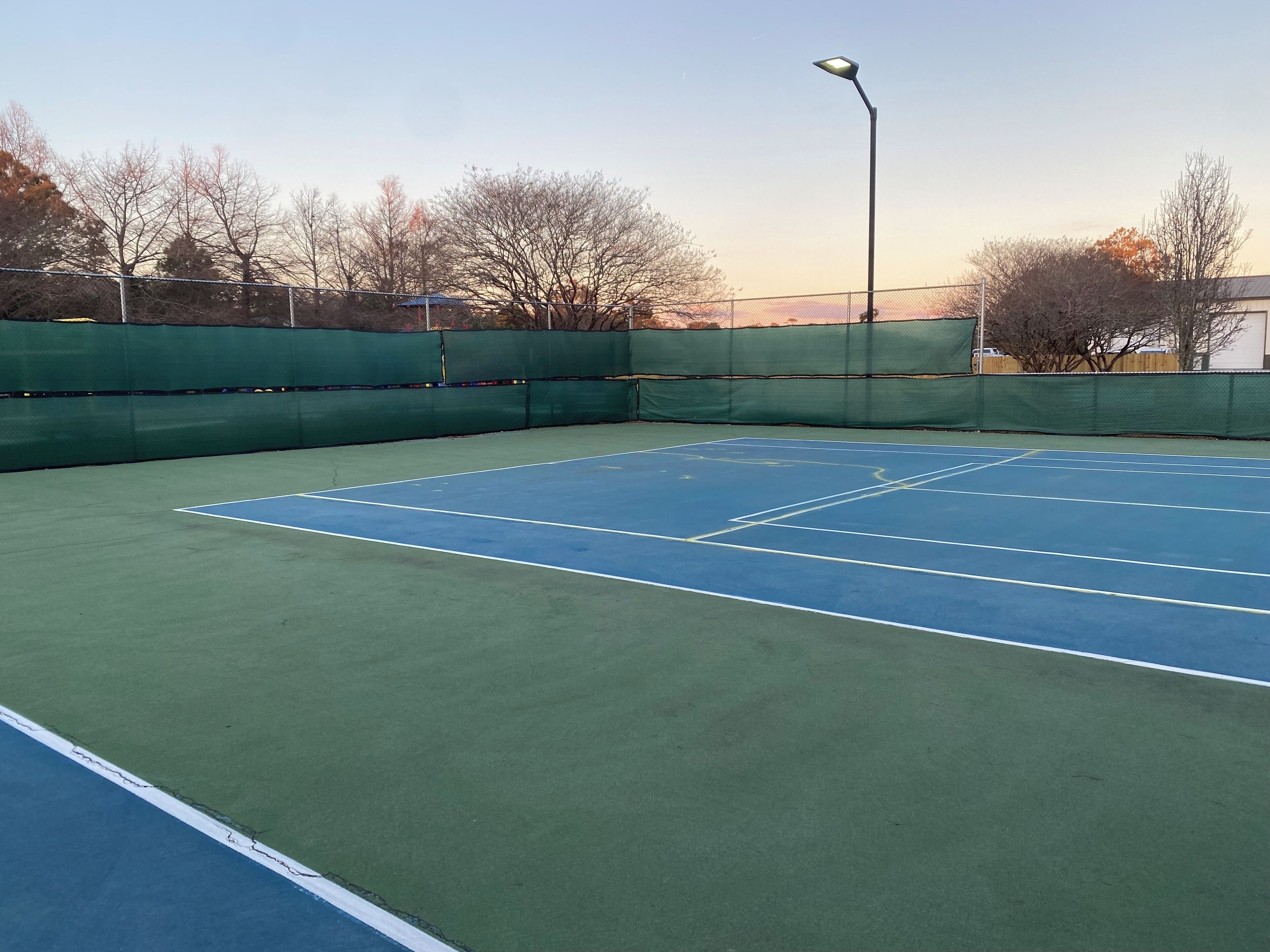 Una cancha de tenis con superficies verde y azul, rodeada por una cerca verde y árboles.