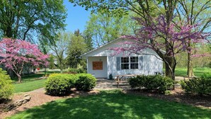 Une petite maison blanche avec un porche, entourée d’une végétation luxuriante et d’arbres en fleurs.