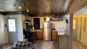Une cuisine moderne avec un réfrigérateur en acier inoxydable, des armoires en bois et un tableau noir où figure une liste de tâches à accomplir. Le salon est agrémenté de chaises à carreaux et une porte porte une pancarte.