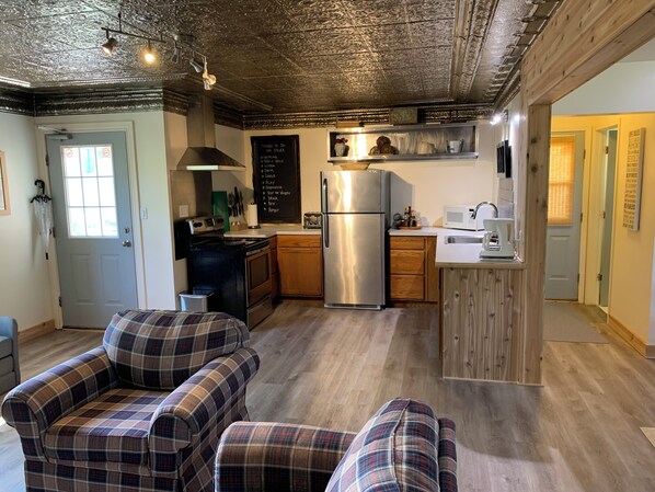 A modern kitchen with a stainless steel refrigerator, wooden cabinets, and a chalkboard with a list of things to do. There are plaid chairs in the living area and a door with a sign.