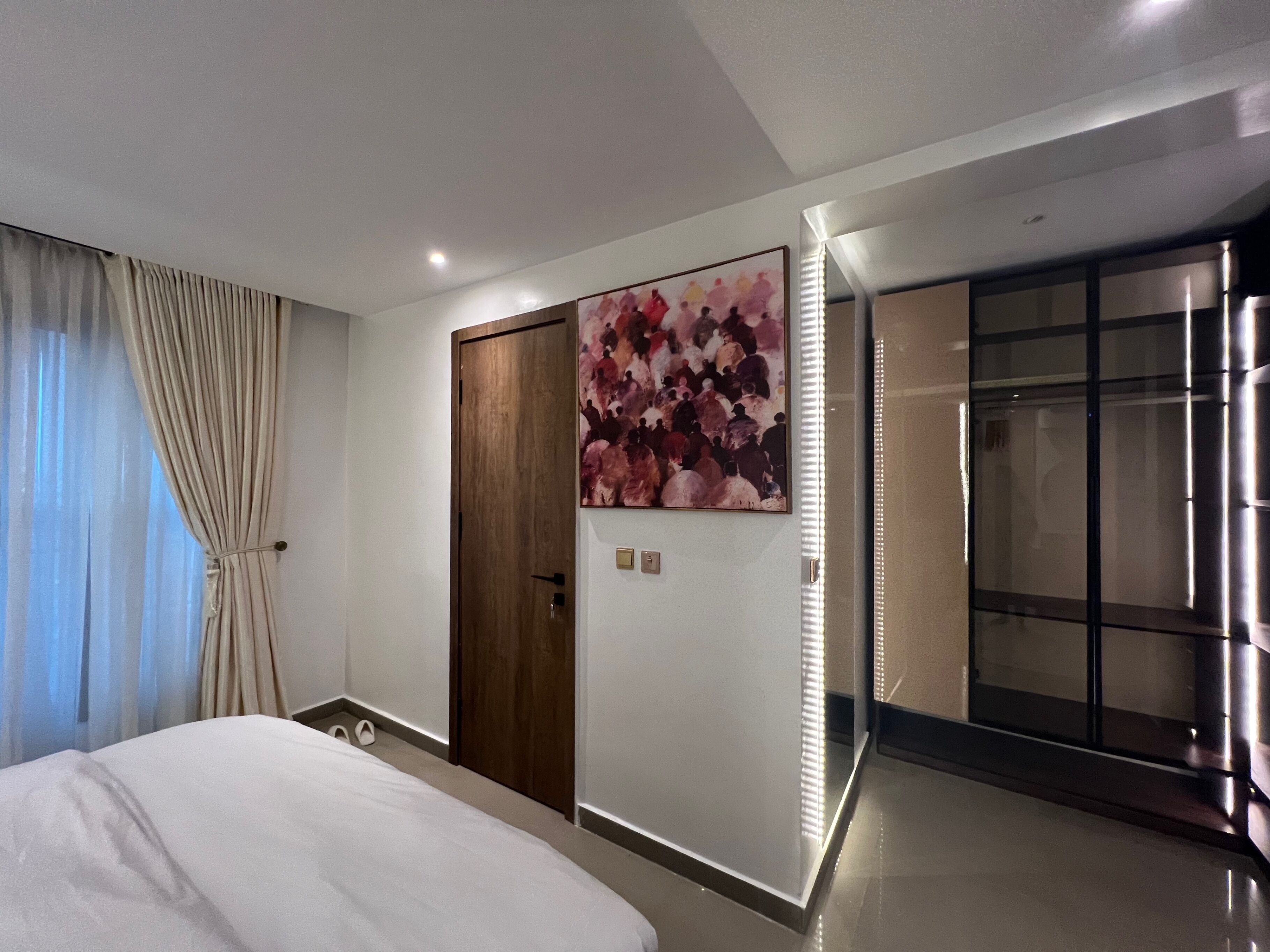 A modern hotel room with a large bed, a wooden wardrobe, a painting on the wall, and a sliding door leading to a bathroom.