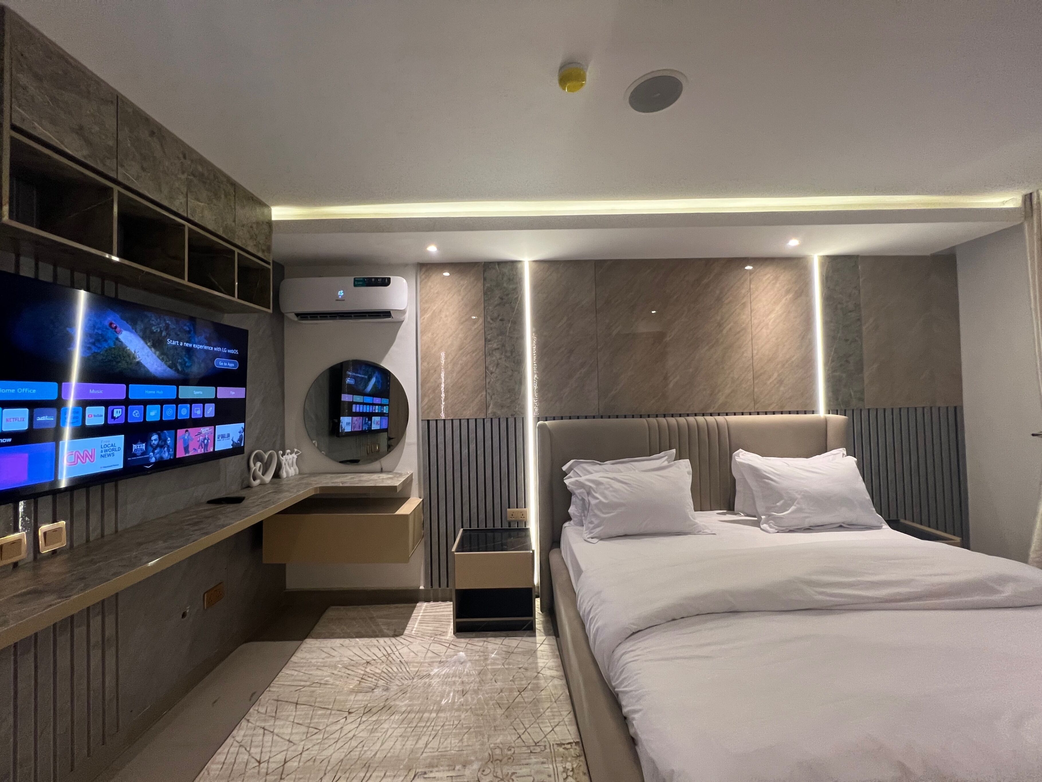 A modern hotel room with a large flat-screen TV mounted on the wall, a round mirror, a bed with white linens, and a built-in air conditioning unit.