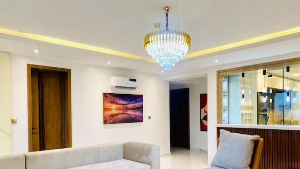 A modern living room with a beige sofa, a wooden chair, a chandelier, and a painting of a sunset.