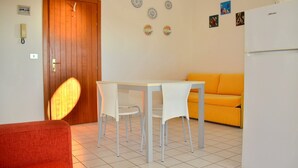 A small, well-lit room with a wooden door, a white table with two chairs, a yellow sofa, and a white refrigerator.
