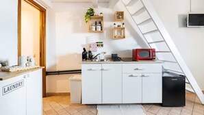 Cozinha compacta com armários brancos, micro-ondas e um pequeno frigorífico. Há uma área de lavandaria à esquerda e uma prateleira de madeira com diversos objetos na parede.