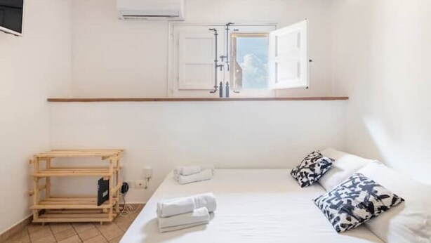 Quarto com cama, janela com vista, prateleira de madeira e ar-condicionado fixado na parede.
