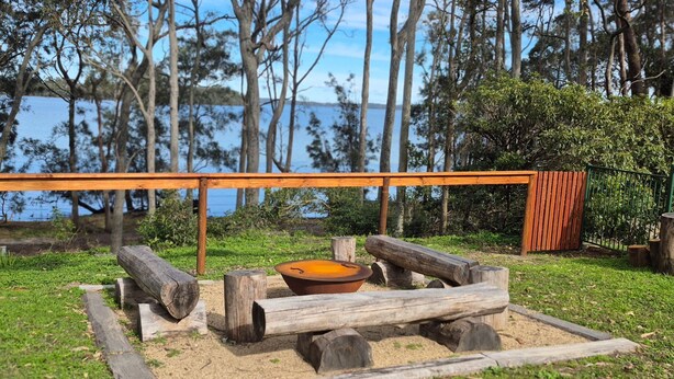 Un brasero de madera con leños y un cuenco, rodeado de un área cubierta de césped con árboles y una valla al fondo.