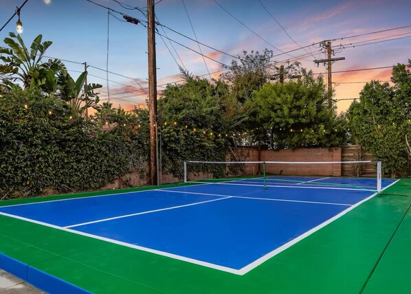 An outdoor tennis court with a green and blue surface, surrounded by dense foliage and a sunset in the background. - Pickleball Disney Resort w/ heated pool &sleeps 12 (Santa Ana)