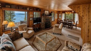A cozy cabin interior with a fireplace, comfortable seating, and a large window overlooking a lake.