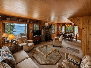 A cozy cabin interior with a stone fireplace, wooden walls, a large window overlooking a lake, and a dining area.