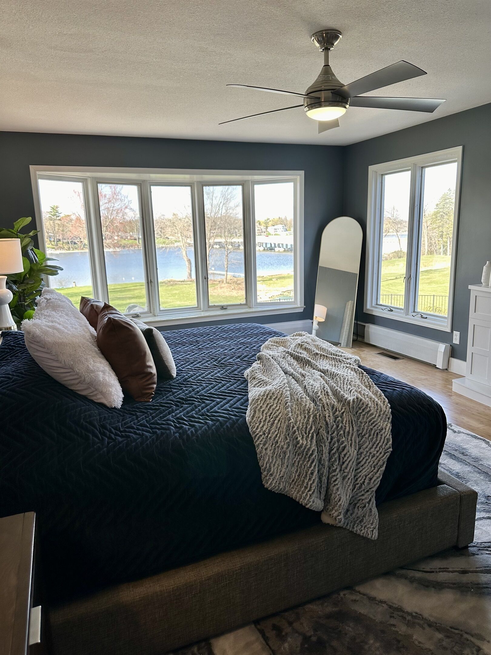 A bedroom with a large bed, a ceiling fan, and a view of a lake through large windows.