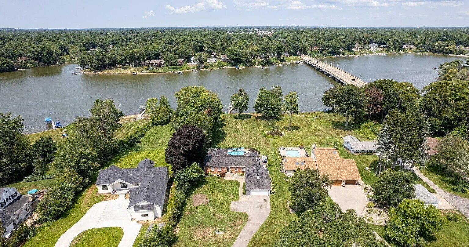 An aerial view of a residential area with a lake, a bridge, and houses with lawns and driveways.