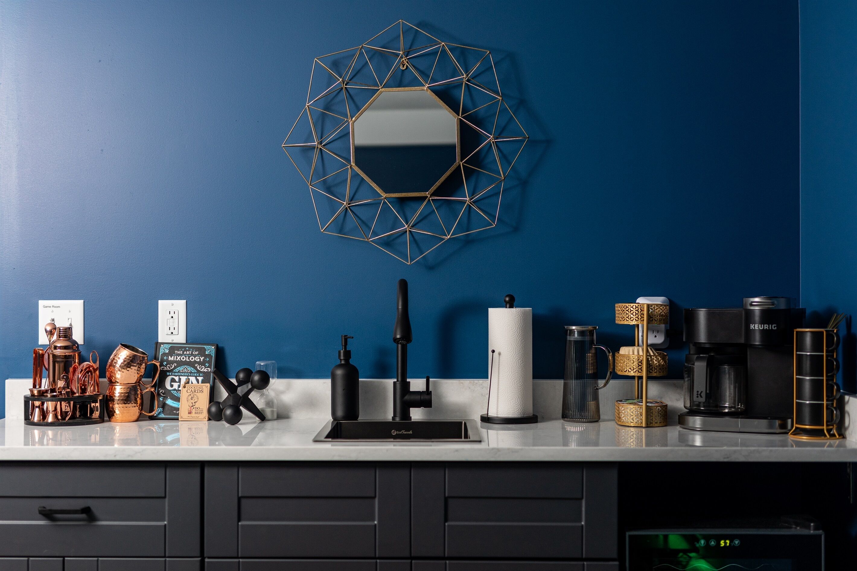 A kitchen with a blue wall, a geometric mirror, a coffee maker, a kettle, and various kitchenware.