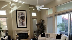 A spacious living room with a fireplace, a ceiling fan, and large windows offering a view of the outdoors. - Beautiful home away from home, nestled in the center of fabulous Indian Ridge CC (Palm Desert)