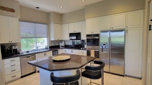 A modern kitchen with white cabinets, a stainless steel refrigerator, a built-in oven, and a large island with bar stools. - Beautiful home away from home, nestled in the center of fabulous Indian Ridge CC (Palm Desert)
