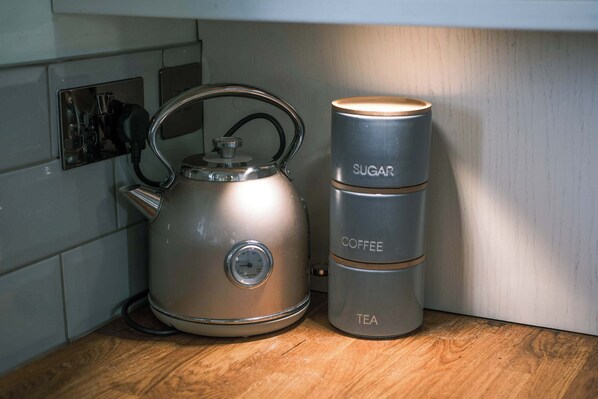 A stainless steel kettle and a stack of containers labeled 'SUGAR,' 'COFFEE,' and 'TEA' on a wooden countertop.