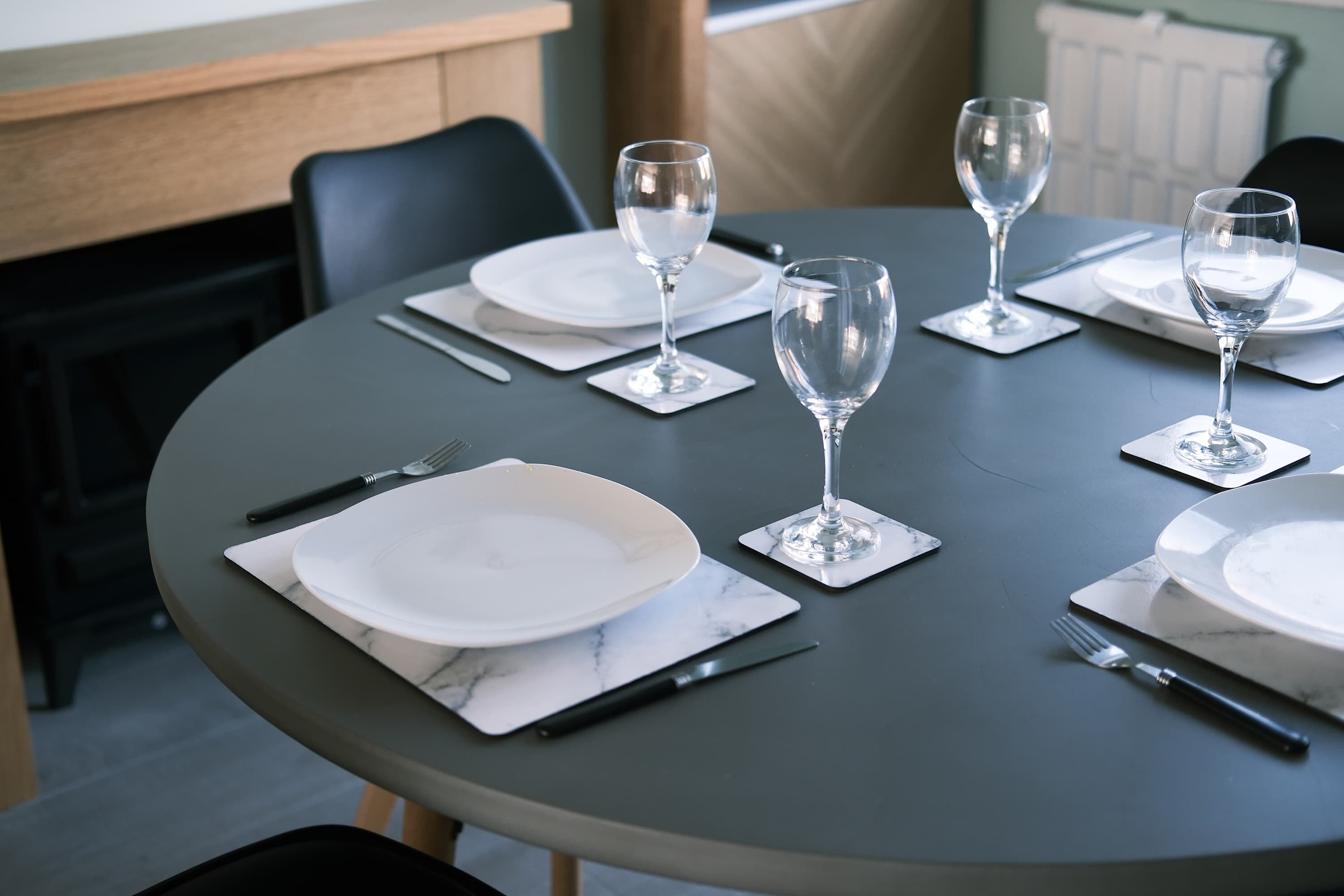 A round dining table with white plates, silverware, and clear wine glasses set for four.