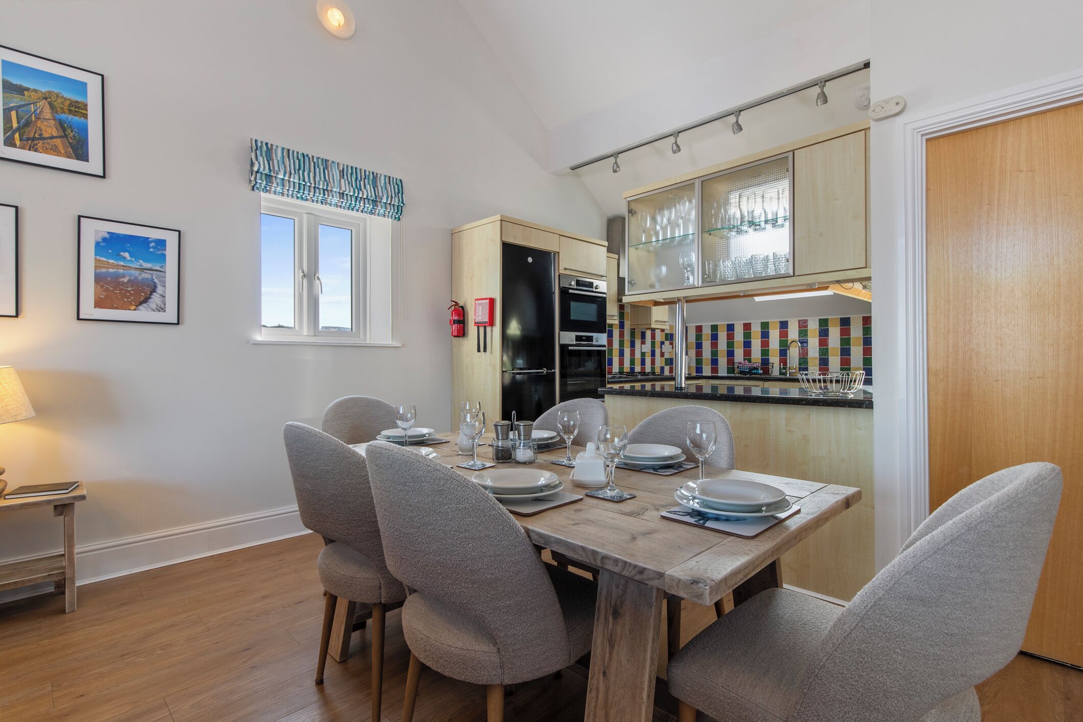 A dining area with a wooden table set for four, surrounded by grey chairs. A kitchen with a tiled backsplash and wooden cabinets is visible in the background.