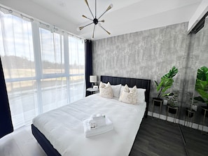 Une chambre à coucher moderne avec un grand lit, un ventilateur de plafond, une table de chevet, une lampe et une vue sur l’extérieur.