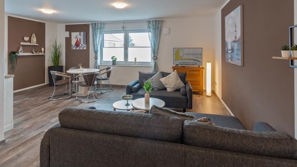 Een moderne woonkamer met een grijze bank, een eettafel met stoelen, een klein bijzettafeltje en een open haard. Er is een raam met gordijnen, een schilderij aan de muur en een plant op de vloer.