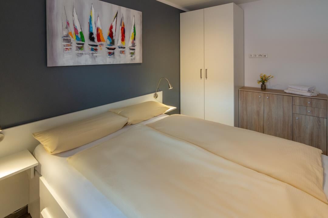 Een slaapkamer met een bed, een nachtkastje, een garderobe en een schilderij van zeilboten aan de muur.